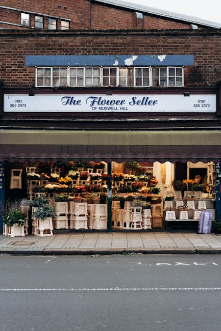 The exterior of 'The Flower Seller' shop on Muswell Hill showcases a vibrant display of floral arrangements through the open storefront. Outside, numerous white wooden plant stands and small tables ho