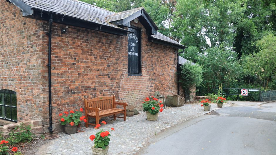 A quaint brick building with a dark wooden sign reading 'PARK FARM & SON MILLHILL' mounted on the wall. In front of the building, there is a wooden bench placed on a cobblestone surface, surrounded by