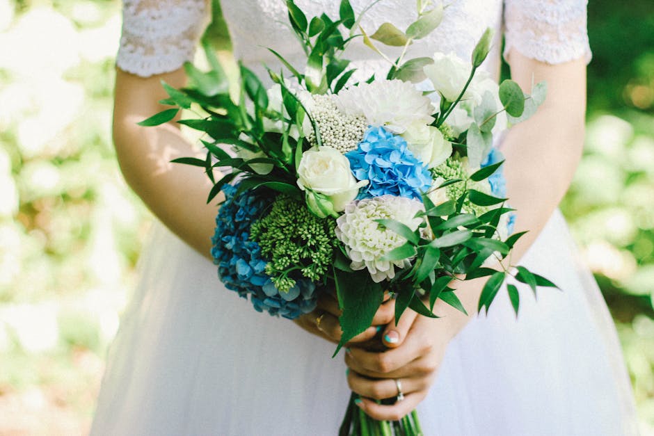 A person in a white dress holding a vibrant floral bouquet, featuring white roses, blue hydrangeas, white carnations, and small green foliage. The bouquet is arranged in a rounded, natural style with